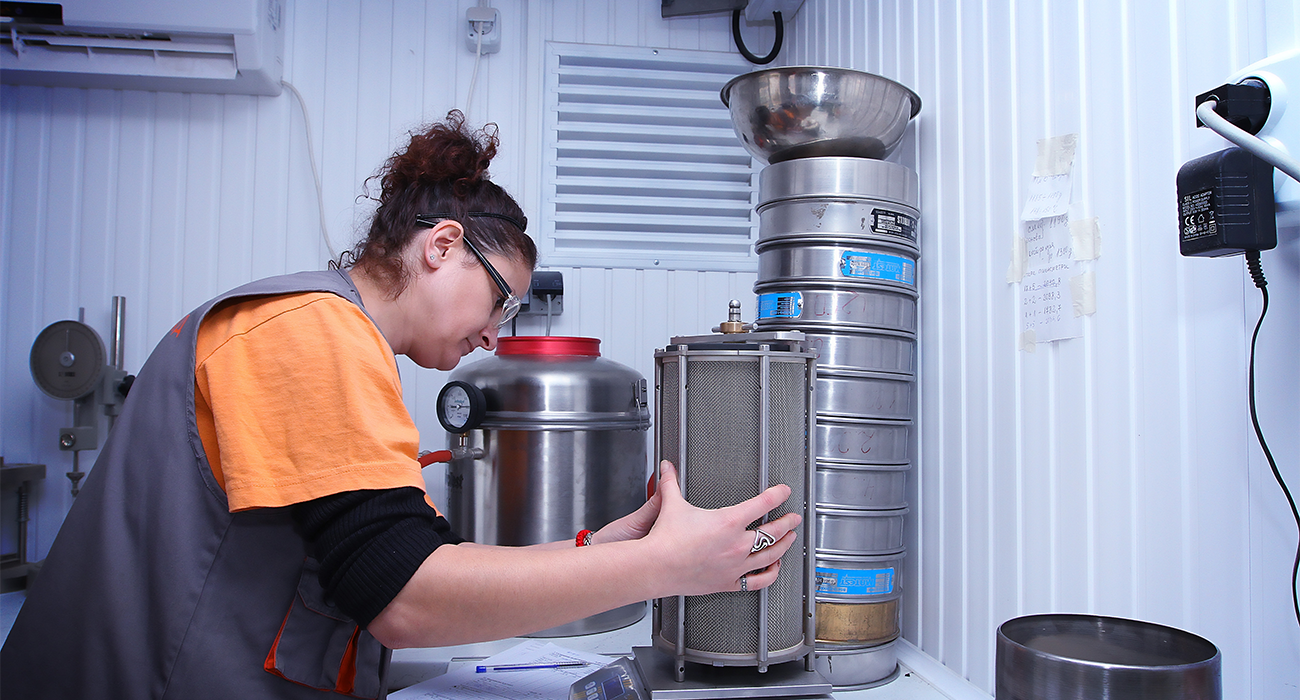 A Road Company employee in work clothes and safety goggles operating a cylindrical sieve in a laboratory.