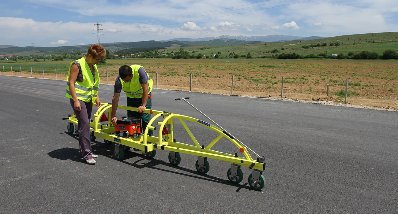 Workers with reflective vests operating a pavement testing device on a paved road.