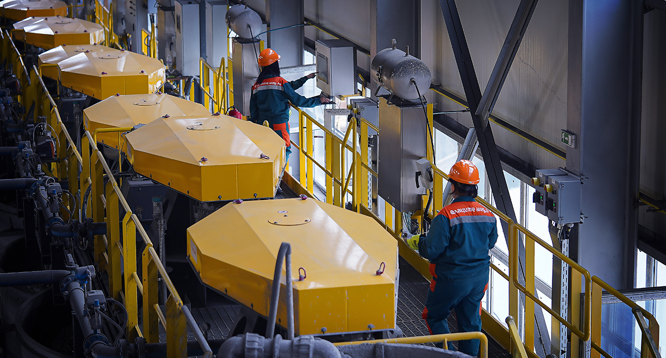 Employees from Ellatzite Med wearing work clothes and safety helmets in a plant with yellow machines.