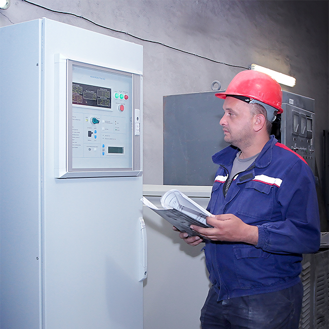 A man in work clothes looking at a wall-mounted digital controller, holding a paper document in his hand.