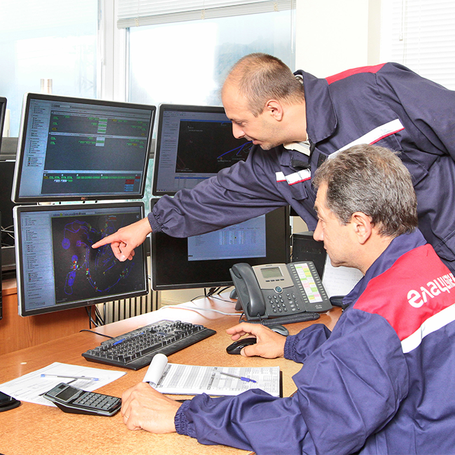 Two Ellatzite-Med employees in work clothes and safety helmets in a control room analyzing data from system software on multiple computer monitors.