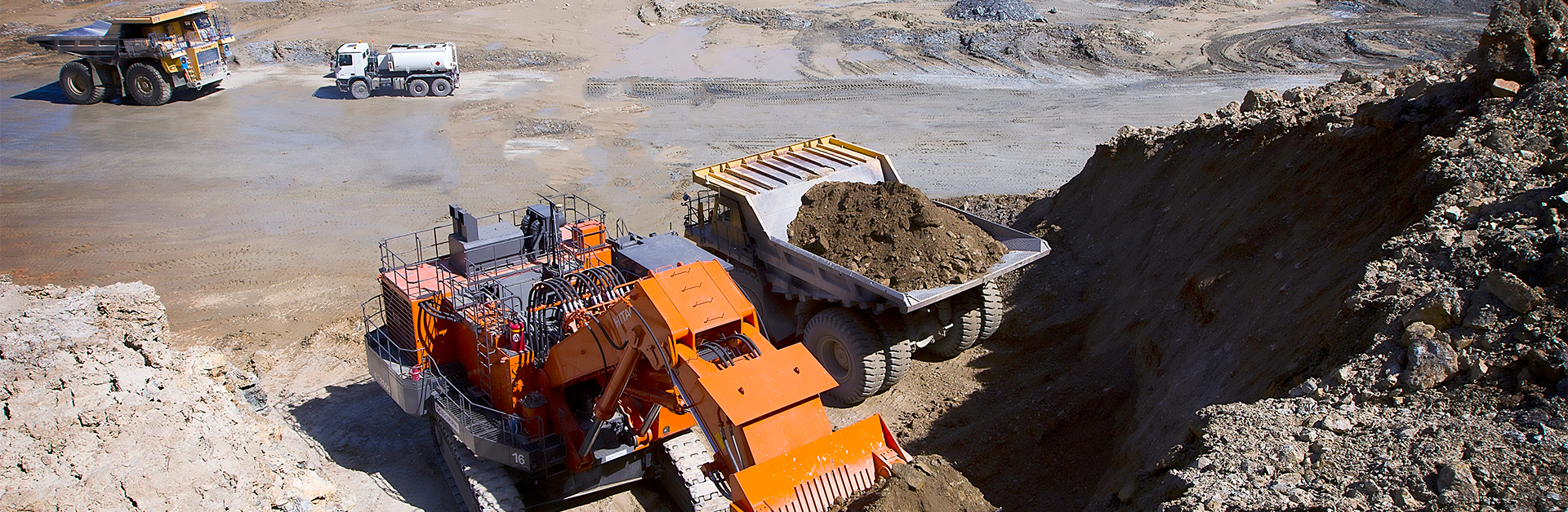 An excavator digging sand and a dump truck next to it carrying the sand. In the background, two more machines for transporting industrial materials are visible.