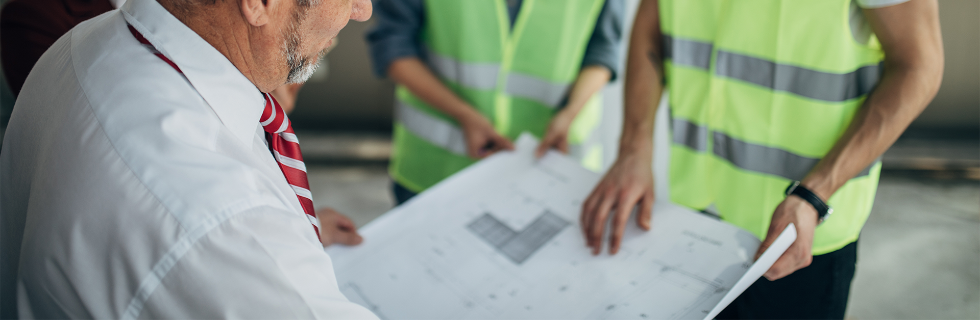 A man in a tie looking at a drawing while two other men in reflective vests point out details on the drawing.