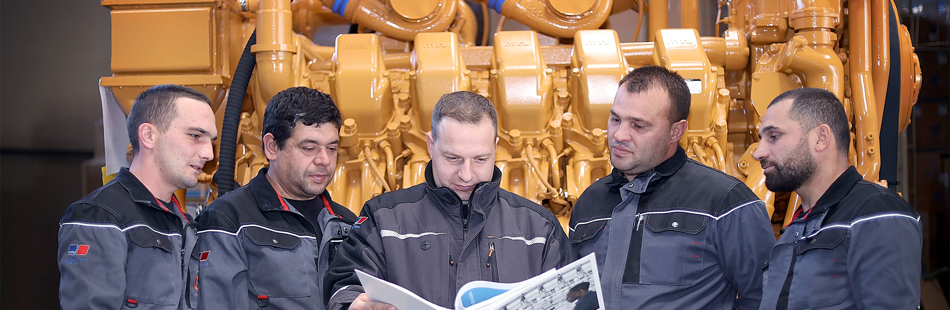 Geotrading employees in work clothes looking at a bulletin in front of a large engine.